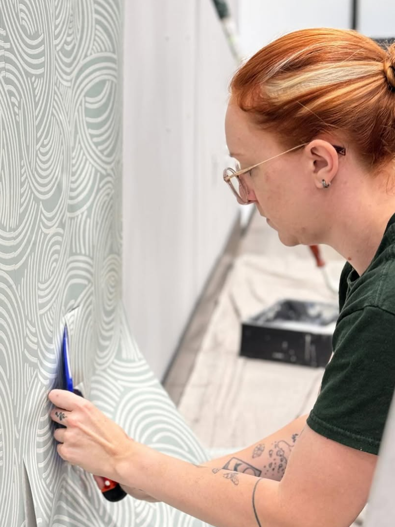 Kirsten Mould of Peridot Designs installing a swirled design wallpaper on an otherwise blank wall. 